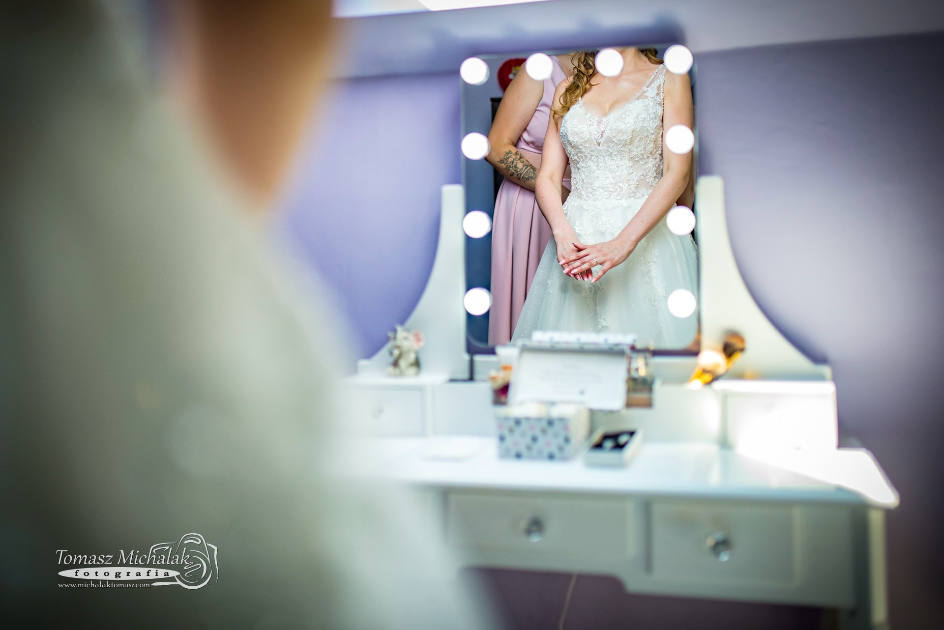 Panna młoda w sukni ślubnej, widoczna w lustrze toaletki z oświetleniem, obok stoi druhna z tatuażem na ramieniu.