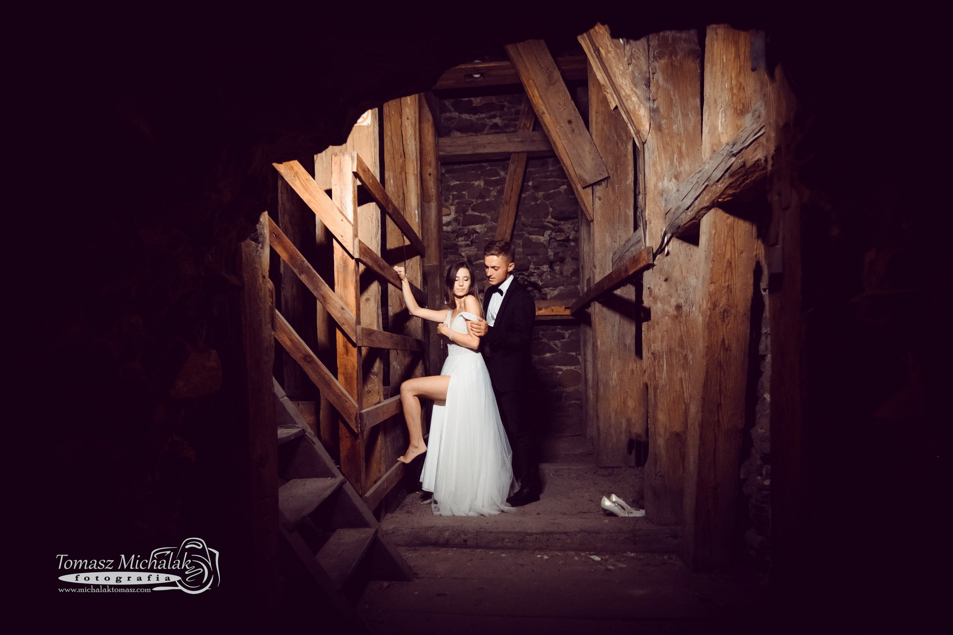 Para młoda w opuszczonej kopalni: panna młoda w białej sukni opiera się o drewniane schody, pan młody w smokingu obejmuje ją, a w tle leżą białe buty.