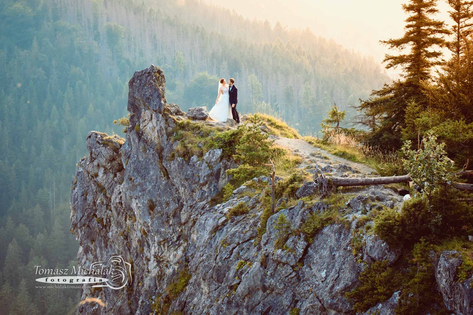 Para młoda w strojach ślubnych stoi na skalistym szczycie góry, w tle zalesione zbocza i delikatna mgła w promieniach zachodzącego słońca.