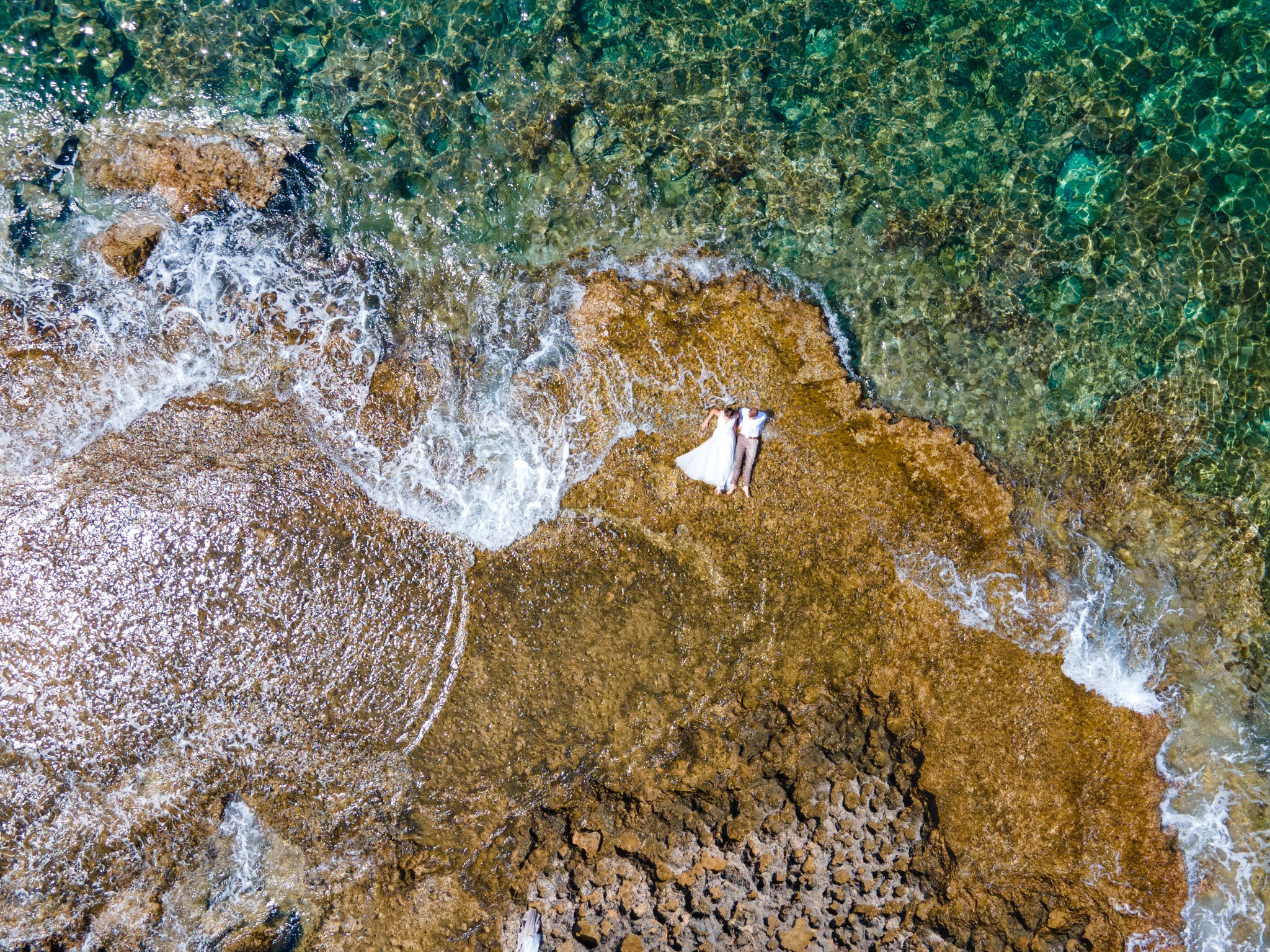 Widok z góry na parę leżącą na skalistym brzegu obok turkusowej wody morskiej, panna młoda w białej sukni.