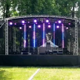 Scena plenerowa z oświetleniem LED w odcieniach fioletu i niebieskiego, konstrukcja aluminiowa, widoczne nagłośnienie i elementy wyposażenia, w tle drzewa i budynek.