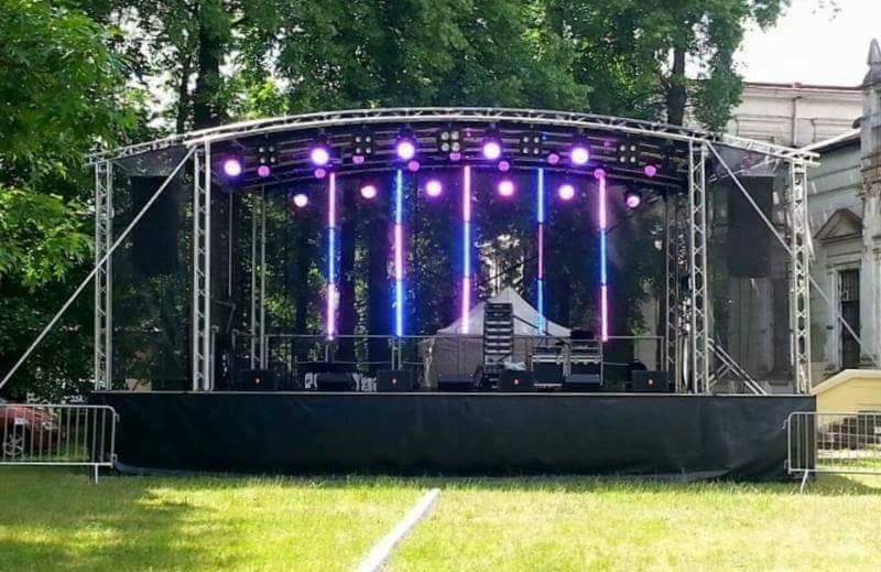 Scena plenerowa z oświetleniem LED w odcieniach fioletu i niebieskiego, konstrukcja aluminiowa, widoczne nagłośnienie i elementy wyposażenia, w tle drzewa i budynek.