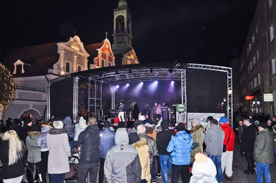 Wieczorny koncert plenerowy na tle zabytkowej architektury, scena oświetlona fioletowymi reflektorami, widoczni muzycy i zgromadzony tłum widzów.