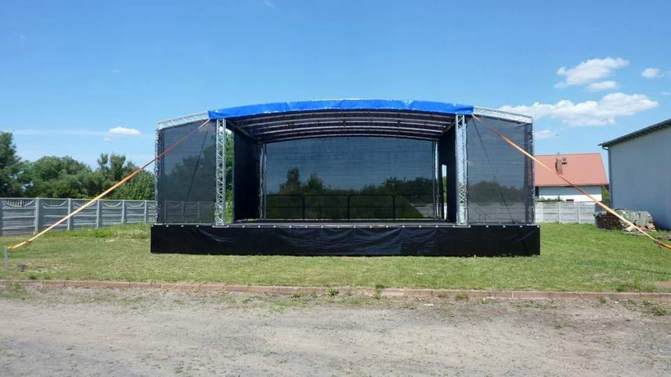 Rozstawiona scena plenerowa z czarnym ekranem i niebieskim zadaszeniem, zabezpieczona pomarańczowymi pasami mocującymi, na trawiastym terenie.