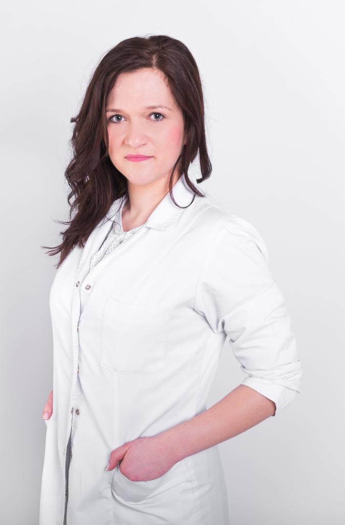 Portret kobiety w białym uniformie medycznym, z ręką w kieszeni, na jasnym tle.