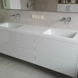 Nowoczesna, minimalistyczna łazienka z podwójną umywalką nablatową, białą szafką pod umywalką z frontami bez uchwytów i bateriami ściennymi, na tle ściany wykończonej płytkami imitującymi falę.