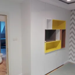 Biała szafa wnękowa z otwartymi p&oacute;łkami w kolorze ż&oacute;łtym, szarym i brązowym na tle ściany w zygzaki, widoczna folia ochronna na podłodze i drabina.