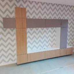 Nowoczesna zabudowa meblowa z elementami drewnianymi i szarymi na tle ściany w zygzaki, widoczne otwory na kable i zabezpieczona podłoga.