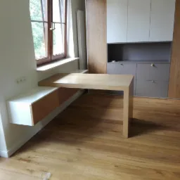 Jasny drewniany blat biurka przymocowany do parapetu okna, z szafką pod spodem, na tle zabudowy meblowej z szafami i szufladami w odcieniach bieli, szarości i drewna.