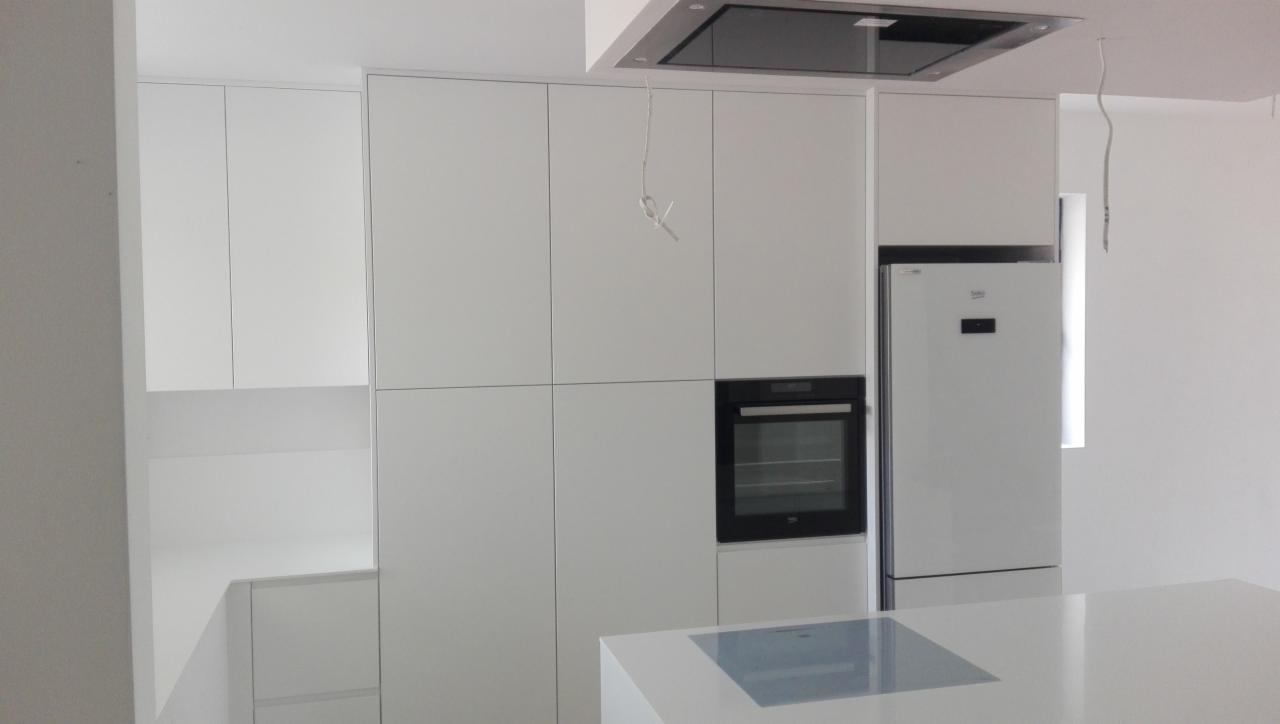 Minimalistyczna, biała zabudowa kuchenna z piekarnikiem i lodówką, widoczne kable wystające z sufitu, blat z wbudowaną płytą indukcyjną.