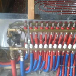 Rozdzielacz ogrzewania podłogowego w metalowej obudowie, z widocznymi czerwonymi i niebieskimi rurami oraz pętlami instalacji w tle, zamontowany między drewnianymi elementami konstrukcyjnymi.