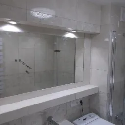 Jasna łazienka z prysznicem za szklaną kabiną, duże lustro z oświetleniem i dekoracyjnymi płytkami z motywem roślinnym, pralka pod blatem.