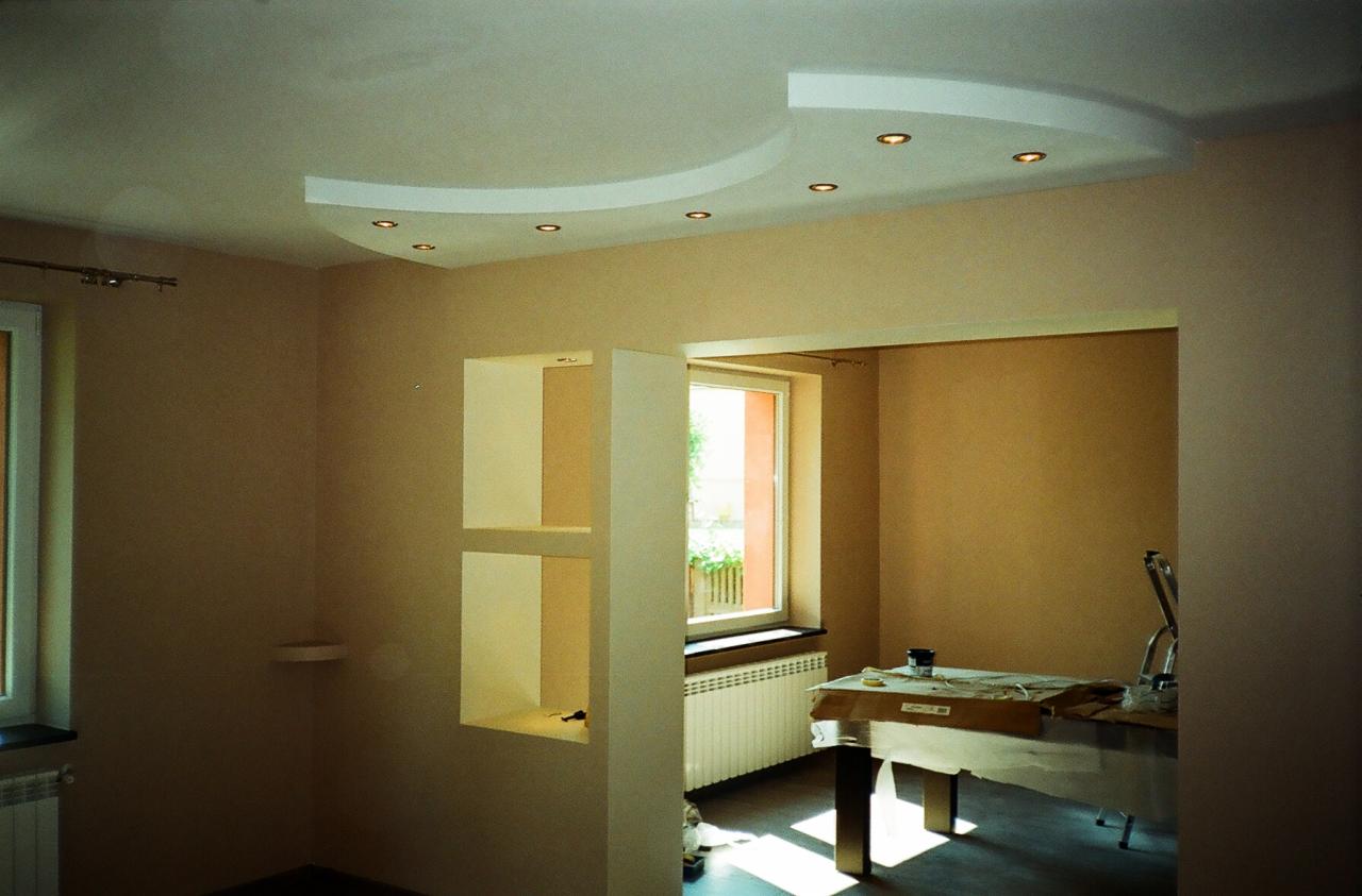 Wykończone wnętrze pokoju z podwieszanym sufitem z oświetleniem punktowym, ścianami w odcieniach beżu i wnękami w ścianie, widok na sąsiednie pomieszczenie z oknem i grzejnikiem.