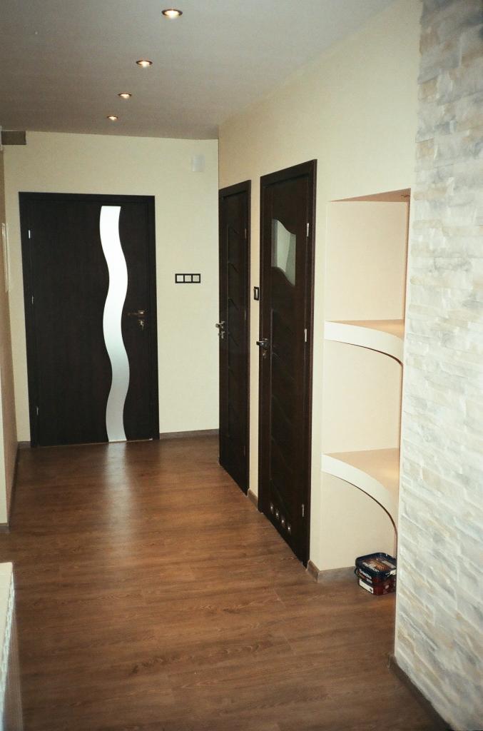 Korytarz po remoncie z panelami podłogowymi imitującymi drewno, dwiema parami ciemnych drzwi (jedne z falistym przeszkleniem), ścianą wyłożoną kamieniem dekoracyjnym i wnęką z półkami.