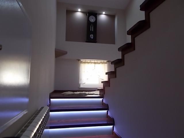 Nowoczesne schody drewniane z podświetleniem LED, prowadzące na antresolę z oknem i dekoracyjnym zegarem ściennym.