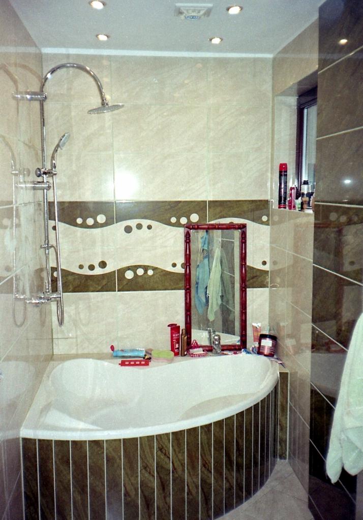 Narożna wanna obudowana płytkami imitującymi drewno, bateria prysznicowa z deszczownicą, dekoracyjne pasy płytek z motywem fal i kropek, lustro w drewnianej ramie, kosmetyki na wannie.