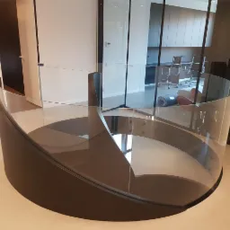 Nowoczesne, zakrzywione schody z czarną podstawą i szklaną balustradą, widok z góry, w tle biuro z szarymi meblami.