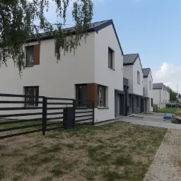 Rząd nowo wybudowanych, minimalistycznych dom&oacute;w jednorodzinnych z czarnymi dachami i ogrodzeniami, widok z perspektywy ulicy, z elementami krajobrazu miejskiego w tle.