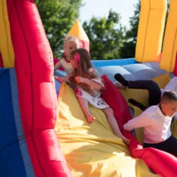 Dzieci w różnym wieku bawią się w dmuchanym zamku w kolorach czerwonym, żółtym i niebieskim, w słoneczny dzień.