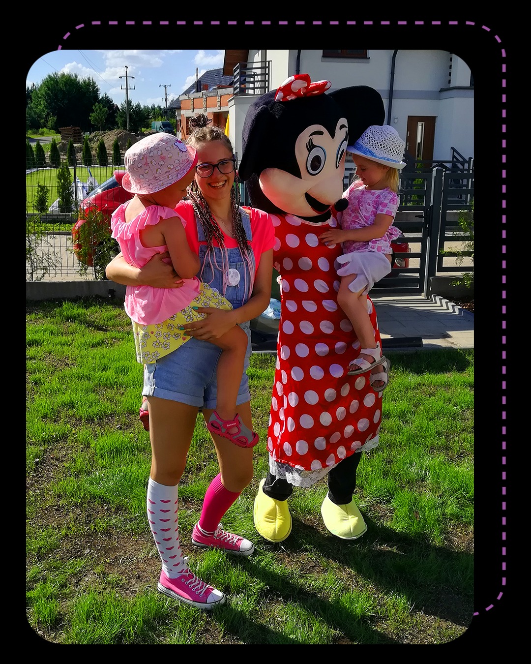 Animator w stroju Myszki Minnie pozuje z dwiema dziewczynkami trzymanymi na rękach przez młodą kobietę w ogrodzie na tle domu.