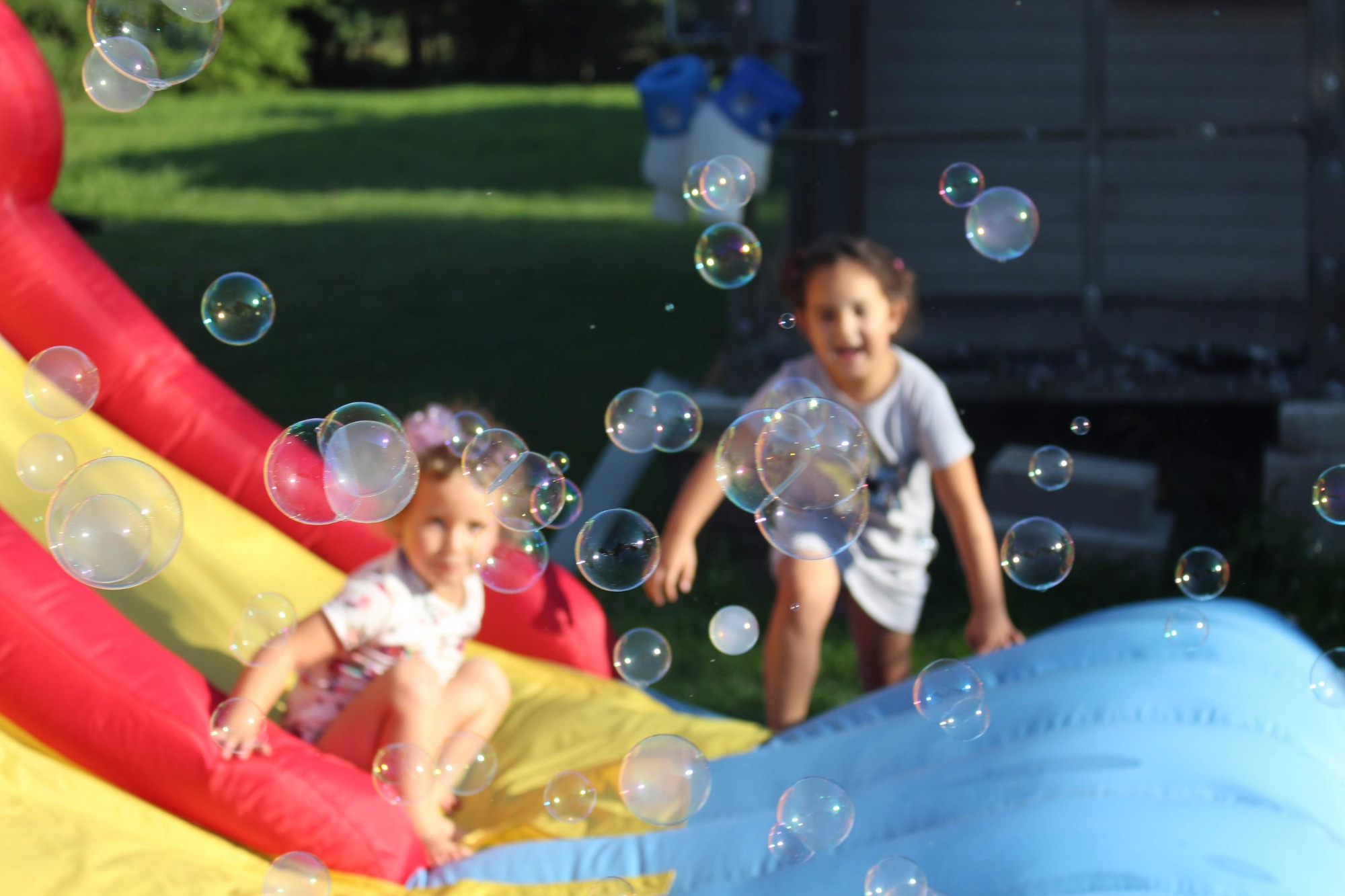 Dwie dziewczynki bawią się na dmuchanym zamku, otoczone unoszącymi się bańkami mydlanymi w słoneczny dzień.