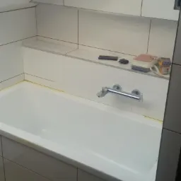 Biała wanna obudowana szarymi płytkami, z chromowaną baterią, białe płytki na ścianach i półce, z widocznymi akcesoriami łazienkowymi oraz niedokładnościami w fugowaniu.