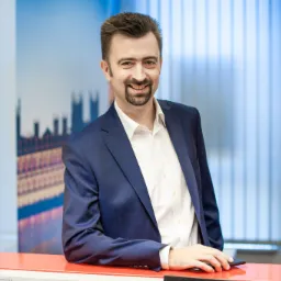 Uśmiechnięty broker ubezpieczeniowy w granatowym garniturze opiera się o pomarańczowe biurko na tle panoramy Londynu.