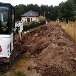 Biała mini koparka Bobcat przygotowuje wykop w ziemi na posesji z domem jednorodzinnym w tle, widoczna linia wykopu wzdłuż ogrodzenia.