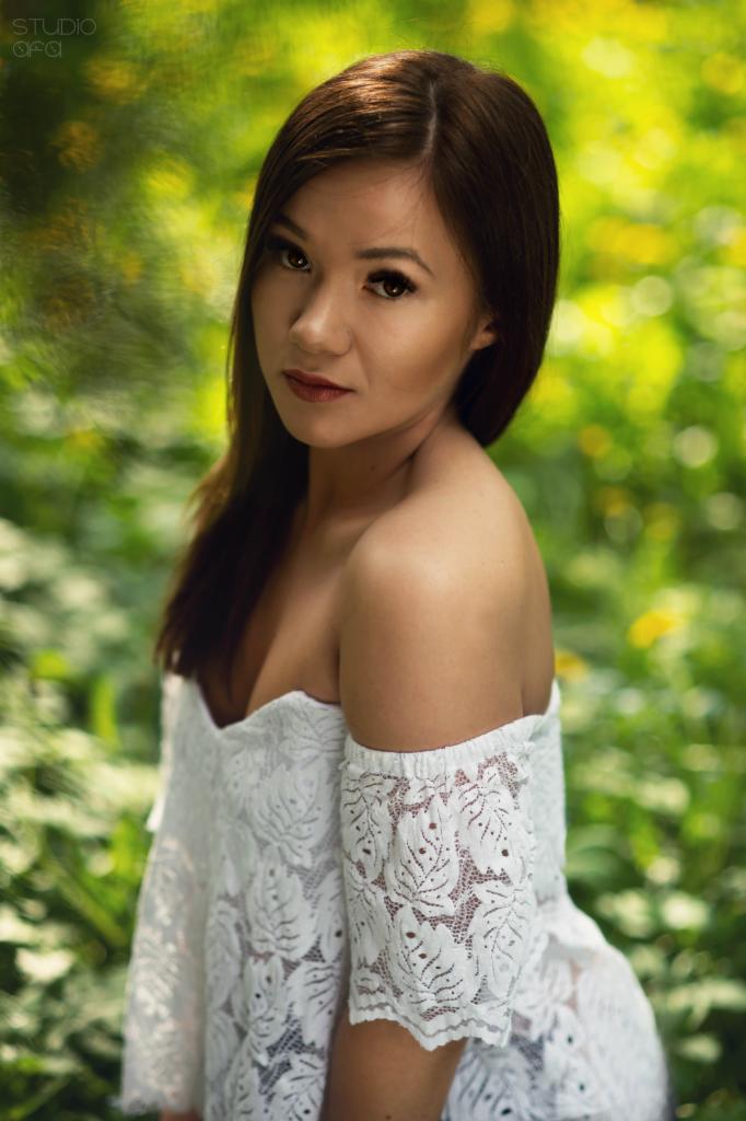 Portret kobiety w białej koronkowej sukience na tle zieleni, z delikatnym rozmyciem tła i naturalnym oświetleniem.