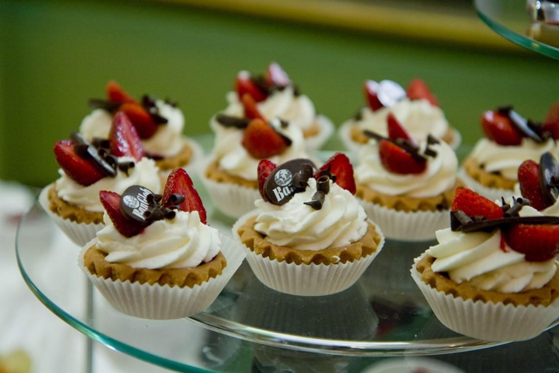 Mini tartaletki z kremem, truskawkami i czekoladowymi dekoracjami na szklanym paterze, widoczne logo cukierni na jednej z nich.