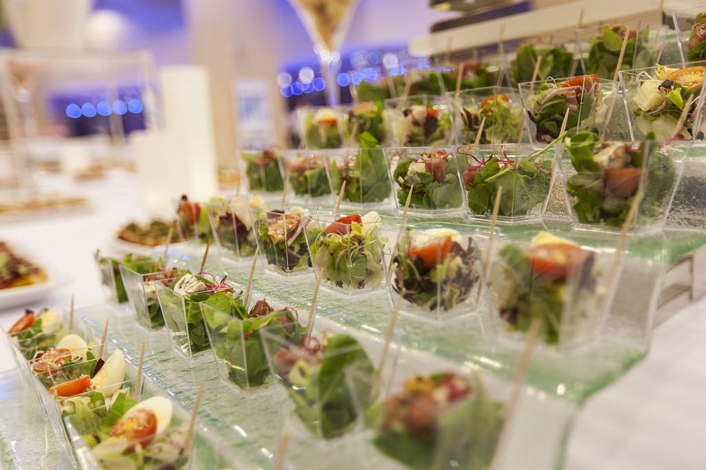 Szereg małych, przezroczystych pojemników z różnorodnymi sałatkami na wielopoziomowej szklanej ekspozycji, z widocznymi elementami dekoracyjnymi w tle.