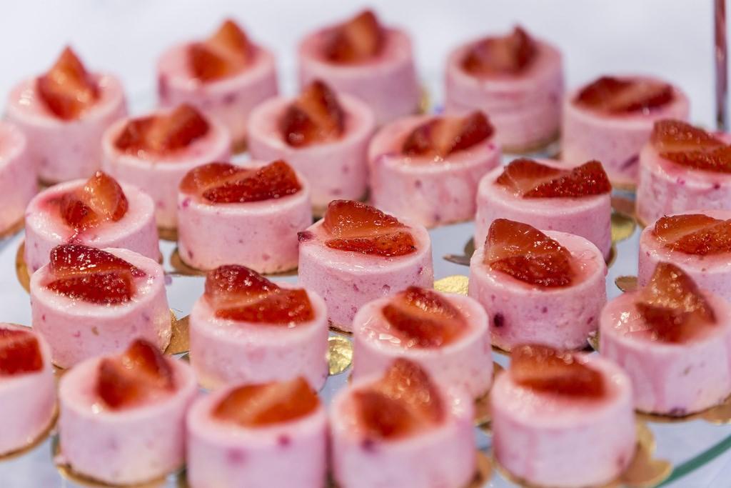 Rząd ciastek musowych o smaku truskawkowym, każdy udekorowany plasterkiem świeżej truskawki na złotym podkładzie, ułożonych na przezroczystej tacy.