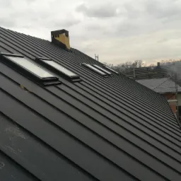 Nowoczesny, czarny dach z blachy na ukośnej konstrukcji z trzema oknami dachowymi, widok z dołu w Lalikach, komin w tle.