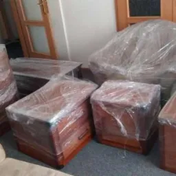 Meble drewniane, w tym krzesło i komody, owinięte folią ochronną przygotowane do transportu w pomieszczeniu z drewnianymi drzwiami w tle.