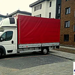 Biały samochód dostawczy z czerwoną plandeką marki Peugeot zaparkowany na parkingu z kostki brukowej przed budynkiem mieszkalnym. Widoczny kontener na śmieci i logo firmy transportowej Zawib Express.