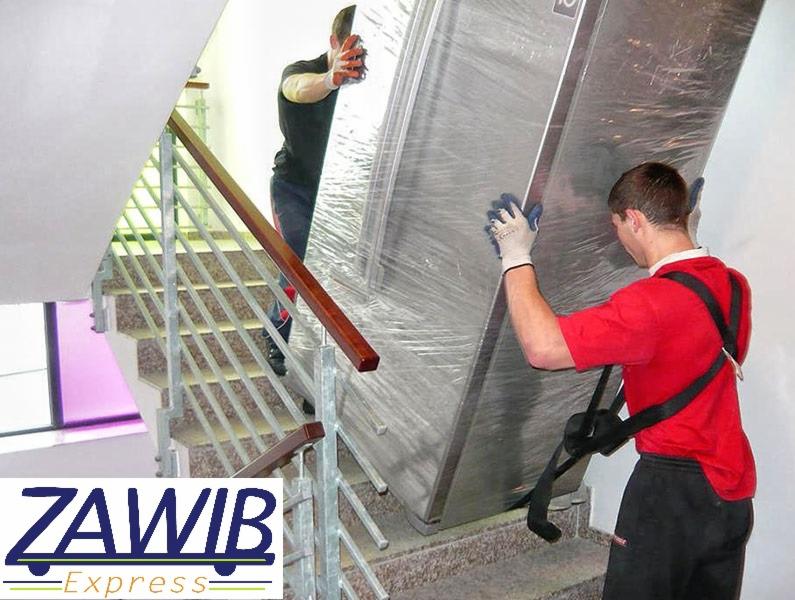 Dwóch mężczyzn wnoszących po schodach dużą, owiniętą folią lodówkę. Jeden mężczyzna z przodu w czerwonej koszulce, drugi z tyłu w ciemnej koszulce. Stalowe schody z drewnianą poręczą i logo firmy...