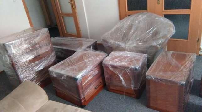 Meble drewniane, w tym krzesło i komody, owinięte folią ochronną przygotowane do transportu w pomieszczeniu z drewnianymi drzwiami w tle.