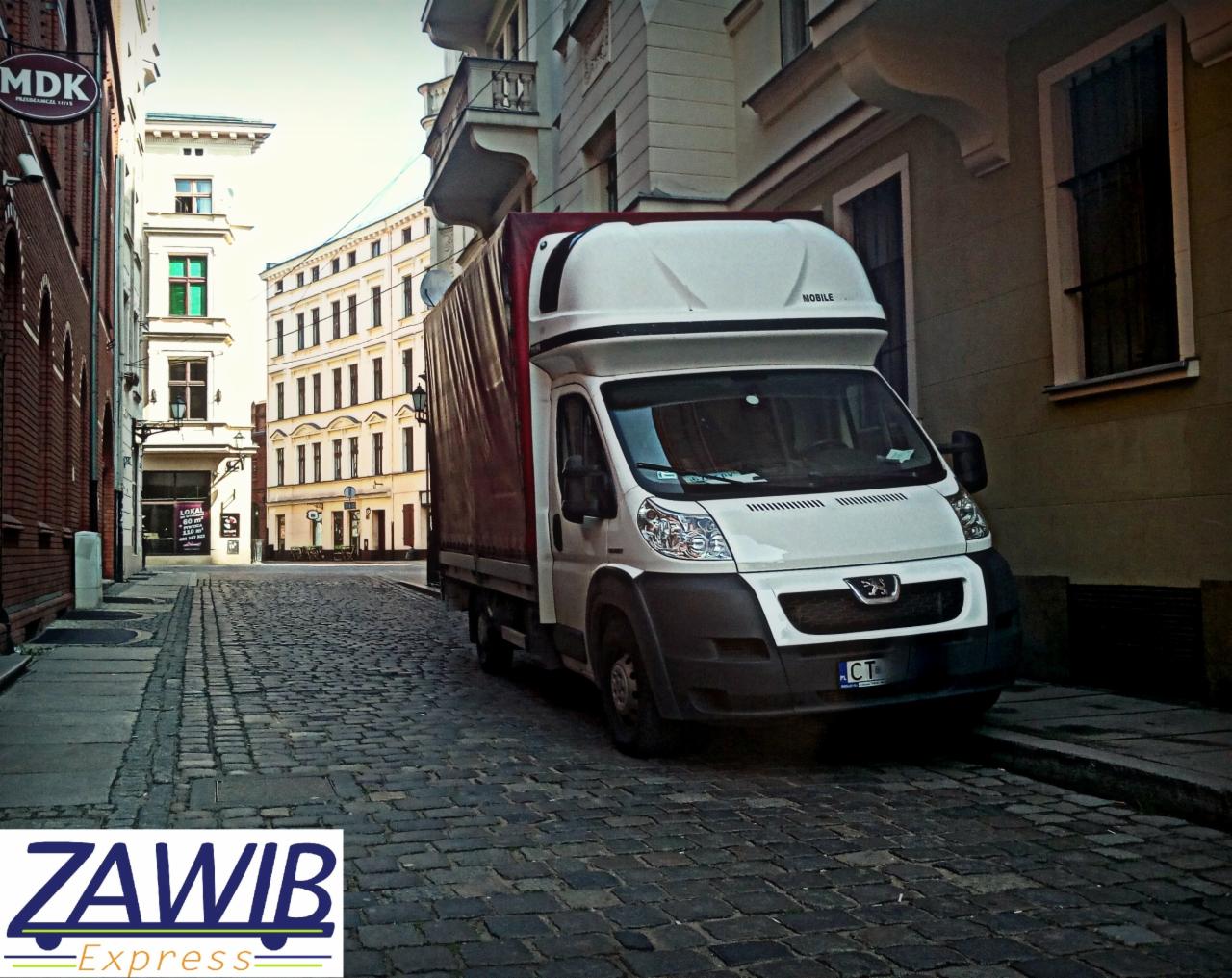 Biały bus dostawczy z plandeką zaparkowany na brukowanej ulicy w Toruniu, w tle zabytkowe kamienice.