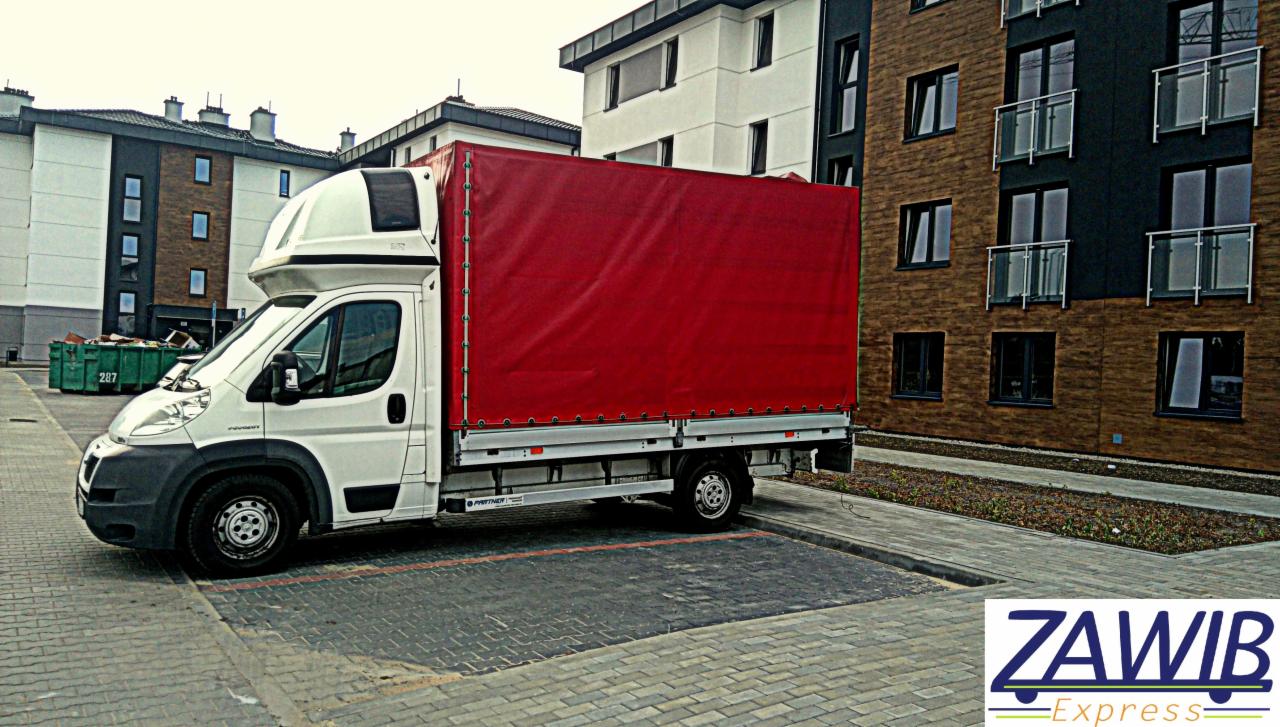 Biały samochód dostawczy z czerwoną plandeką marki Peugeot zaparkowany na parkingu z kostki brukowej przed budynkiem mieszkalnym. Widoczny kontener na śmieci i logo firmy transportowej Zawib Express.