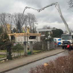 Solutionis Marek Skałbania - Budowa domu jednorodzinnego w trakcie wznoszenia ścian z betonu komórkowego. Na górze konstrukcji drewniany szalunek. Na ulicy stoi betoniarka z pompą do betonu.