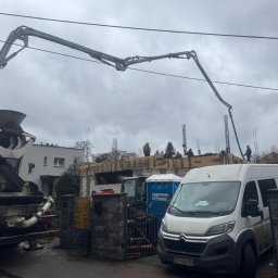 Solutionis Marek Skałbania - Betonomieszarka z wysięgnikiem wylewa beton na budowę domu jednorodzinnego. Widoczni pracownicy w kaskach i biały bus. Pochmurne niebo w tle.