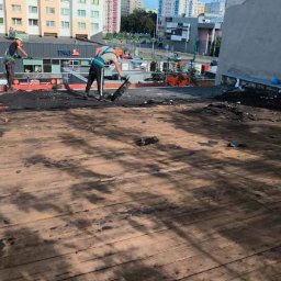 Solutionis Marek Skałbania - Pracownicy w uprzężach podczas prac na płaskim dachu budynku, prawdopodobnie związanych z jego uszczelnianiem lub naprawą. Widoczne narzędzia i materiały budowlane.