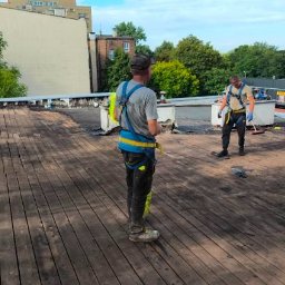 Solutionis Marek Skałbania - Pracownicy w uprzężach na drewnianym dachu podczas prac remontowych. Jeden z nich zamiata. Widok z perspektywy osoby stojącej za nimi.