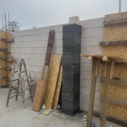 Solutionis Marek Skałbania - Wznoszenie komina z szarej bloczków na budowie. Widoczne białe bloczki ściany, drewniane szalunki, drabina i deski. Surowy stan, etap prac murarskich.