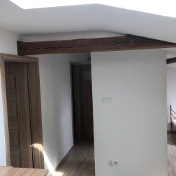 Solutionis Marek Skałbania - Jasne wnętrze po remoncie: drewniane belki stropowe, panele podłogowe imitujące drewno, białe ściany, drzwi z okleiną drewnopodobną. Widok na korytarz.