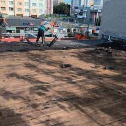 Solutionis Marek Skałbania - Praca na dachu budynku: dwóch robotników w uprzężach usuwa starą papę z drewnianego poszycia, w tle zabudowa miejska i budynek z logo ING.