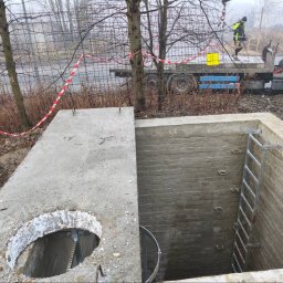 Solutionis Marek Skałbania - Otwarty betonowy właz studzienki kanalizacyjnej z drabinką wewnątrz, częściowo zakryty pokrywą z otworem. W tle ciężarówka z dźwigiem i pracownik w kamizelce.