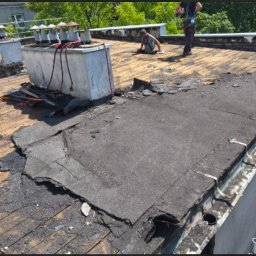 Solutionis Marek Skałbania - Uszkodzony dach budynku z widocznymi pracownikami w trakcie naprawy. Fragmenty starej papy i narzędzia na drewnianej konstrukcji dachu.