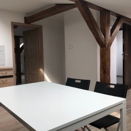 Solutionis Marek Skałbania - Jasne wnętrze z drewnianymi belkami stropowymi i białym stołem. Widoczne drzwi i fragment zabudowy kuchennej. Minimalistyczny design z elementami rustykalnymi.