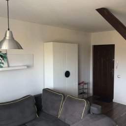 Solutionis Marek Skałbania - Jasne wnętrze mieszkania z szarą sofą z żółtą lamówką, białą szafą i metalową lampą wiszącą. Widoczna drewniana belka stropowa i drzwi wejściowe.